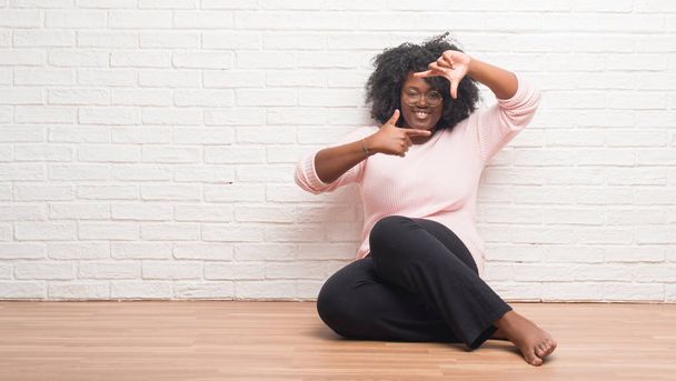 Mujer afroamericana joven sentada en el suelo en casa sonriendo haciendo marco con las manos y los dedos con la cara feliz. Concepto de creatividad y fotografía
. - Foto, imagen