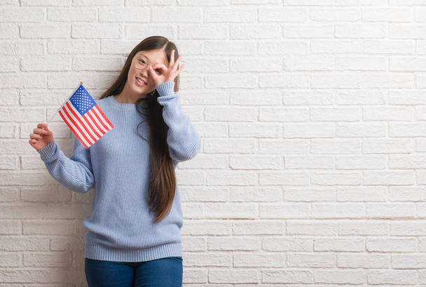 Молода Китайська жінка, що над цегляна стіна тримає прапор Америки з щасливим обличчям, посміхаючись, роблячи ОК знак рукою на-віч, переглядаючи пальці - Фото, зображення