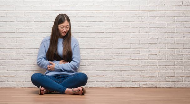Jonge Chinese vrouw zittend op de vloer over bakstenen muur met hand op de maag omdat misselijkheid, pijnlijke ziekte onwel. Ache concept. - Foto, afbeelding