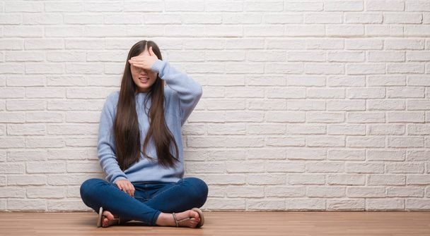 Jeune femme chinoise assise sur le sol au-dessus d'un mur de briques souriant et riant avec la main sur le visage couvrant les yeux pour la surprise. Concept aveugle
. - Photo, image