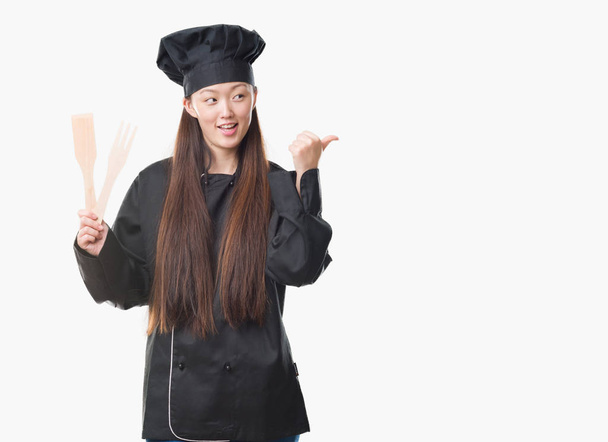 Jeune femme chinoise sur fond isolé portant uniforme de chef pointant et montrant avec le pouce sur le côté avec sourire heureux visage
 - Photo, image
