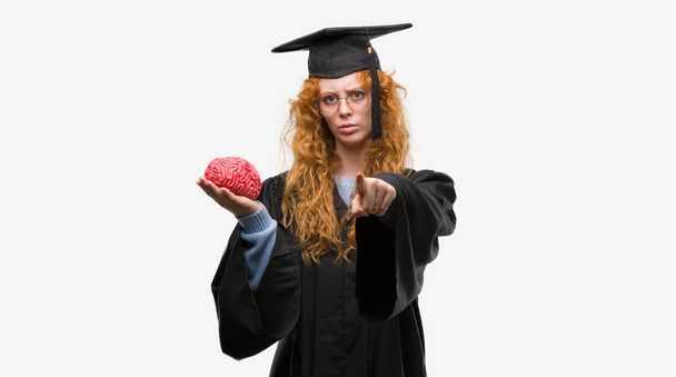 Nuori punapää opiskelija nainen yllään valmistui yhtenäinen tilalla aivot sormella kameraan ja sinulle, käsi merkki, positiivinen ja luottavainen ele edestä
 - Valokuva, kuva