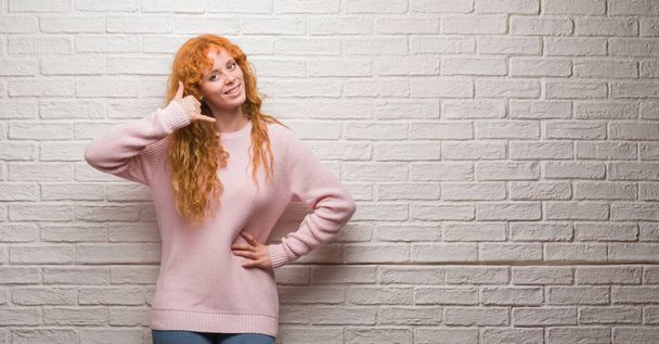 Young redhead woman standing over brick wall smiling doing phone gesture with hand and fingers like talking on the telephone. Communicating concepts. - Φωτογραφία, εικόνα