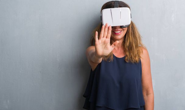 Middle age hispanic woman standing over grey grunge wall wearing virtual reality glasses with open hand doing stop sign with serious and confident expression, defense gesture - Foto, imagen