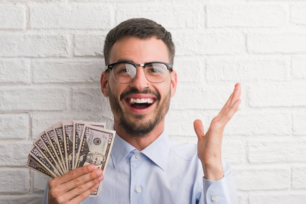 junger Hipster-Geschäftsmann hält Dollars sehr glücklich und aufgeregt in der Hand, Siegerausdruck feiert den Sieg schreiend mit breitem Lächeln und erhobenen Händen - Foto, Bild