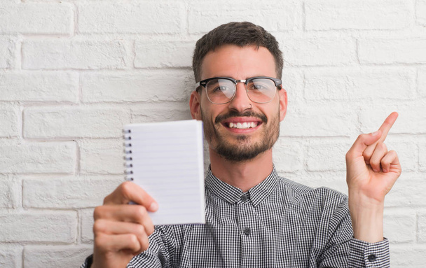 junger erwachsener Mann mit Notizbuch über weißer Ziegelwand stehend sehr glücklich mit Hand und Finger zur Seite zeigend - Foto, Bild