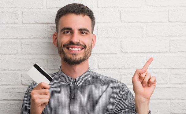 Nuori aikuinen mies tiiliseinän yli pitäen luottokorttia erittäin onnellinen osoittaen kädellä ja sormella sivulle
 - Valokuva, kuva