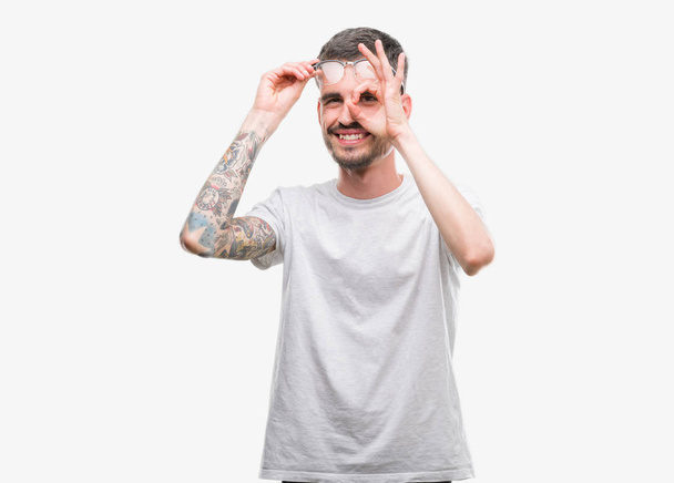 Joven hombre adulto tatuado haciendo buen gesto con la mano sonriendo, los ojos mirando a través de los dedos con la cara feliz
. - Foto, Imagen