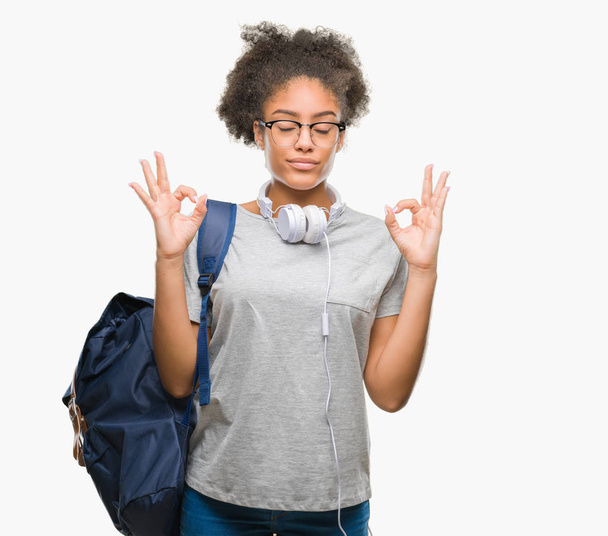 若いアフロ ・ アメリカ人学生女性隔離された背景にヘッドフォンとバックパックを着てリラックスして瞑想の指ジェスチャーをやって閉じた目を浮かべてします。ヨガの概念. - 写真・画像