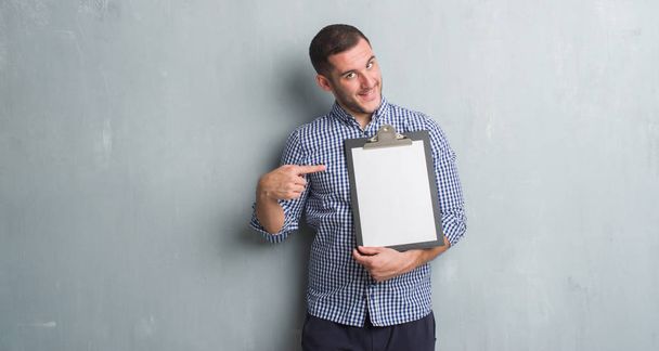 Young caucasian man over grey grunge wall holding clipboard very happy pointing with hand and finger - Photo, Image