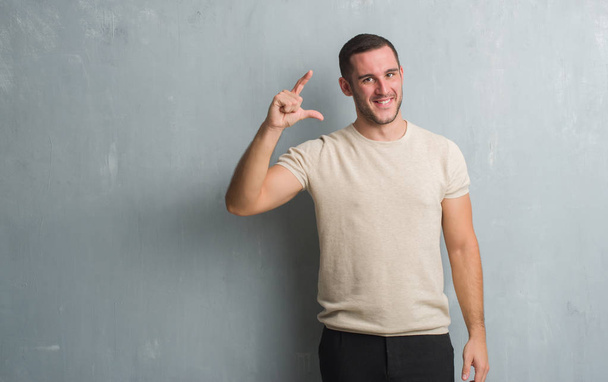 Молодой кавказский мужчина над серой гранж-стеной улыбается и уверенно жестикулирует рукой, делая указатель размера пальцами, глядя в камеру. Концепция измерения
. - Фото, изображение