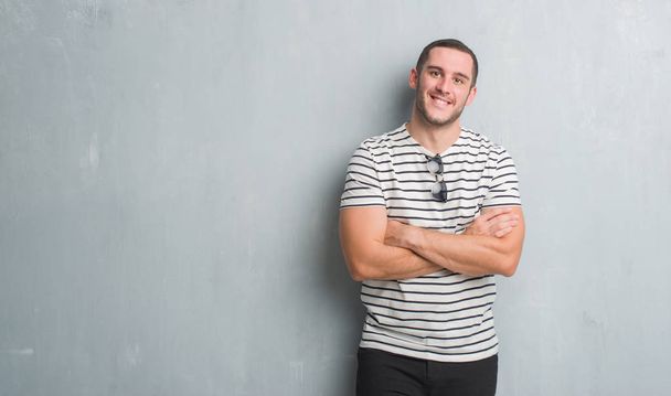 Homem caucasiano jovem sobre parede grunge cinza rosto feliz sorrindo com braços cruzados olhando para a câmera. Pessoa positiva
. - Foto, Imagem