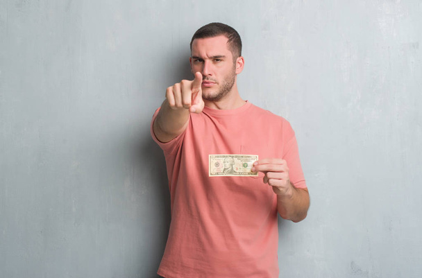 Молодий кавказький чоловік над сірою гранжевою стіною, що показує десять доларів, вказуючи пальцем на камеру і вам, знак руки, позитивний і впевнений жест спереду
 - Фото, зображення