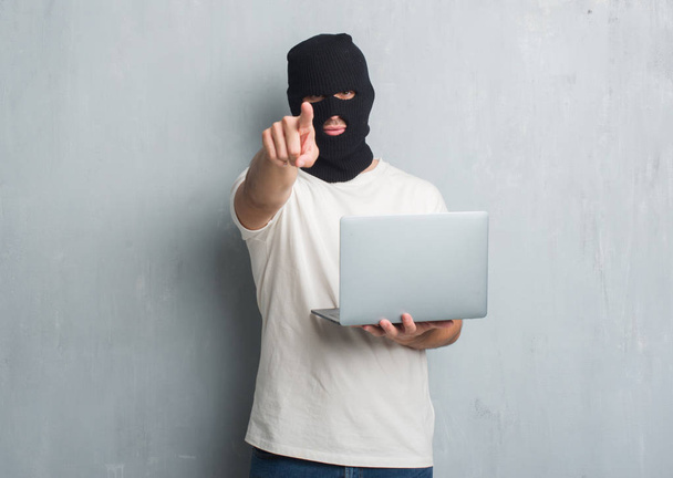 Nuori valkoihoinen hakkeri mies yli harmaa grunge seinä tekee tietoverkkohyökkäys käyttäen kannettava tietokone sormella kameraan ja sinulle, käsi merkki, positiivinen ja luottavainen ele edestä
 - Valokuva, kuva