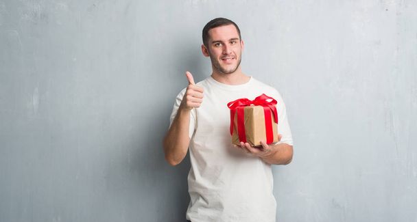 Young caucasian man over grey grunge wall holding a present happy with big smile doing ok sign, thumb up with fingers, excellent sign - Foto, Imagen