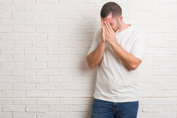 Молодий кавказький чоловік стоїть над білою цегляною стіною з сумним виразом, що покриває обличчя руками під час плачу. Концепція депресії
. - Фото, зображення