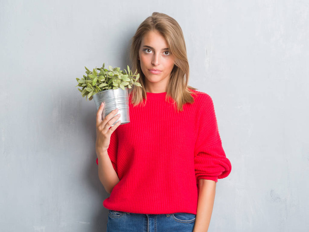 Beautiful young woman over grunge grey wall holding plant pot with a confident expression on smart face thinking serious - Photo, Image