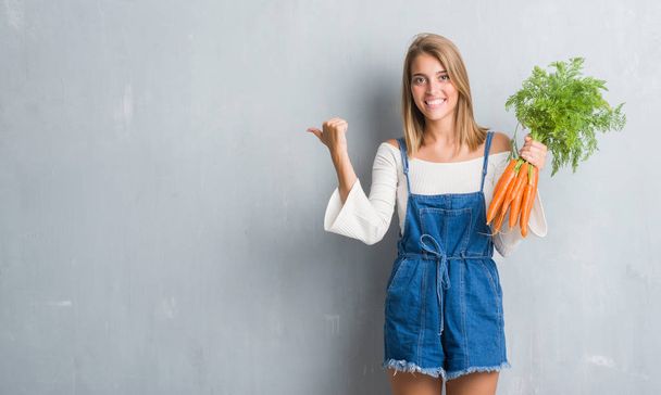 Beautiful young woman over grunge grey wall holding fresh carrots pointing and showing with thumb up to the side with happy face smiling - Photo, Image