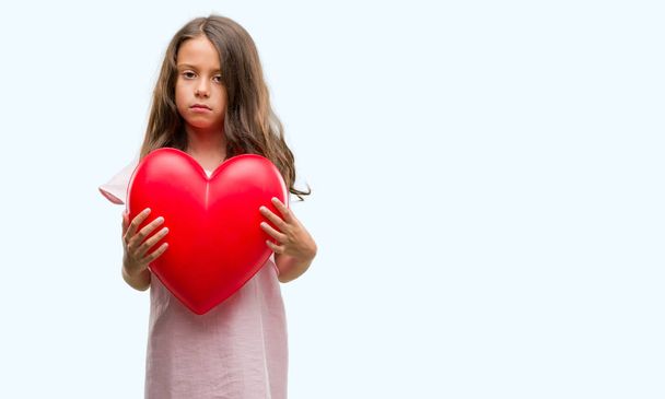 Brunette hispanic girl holding red heart with a confident expression on smart face thinking serious - Foto, imagen