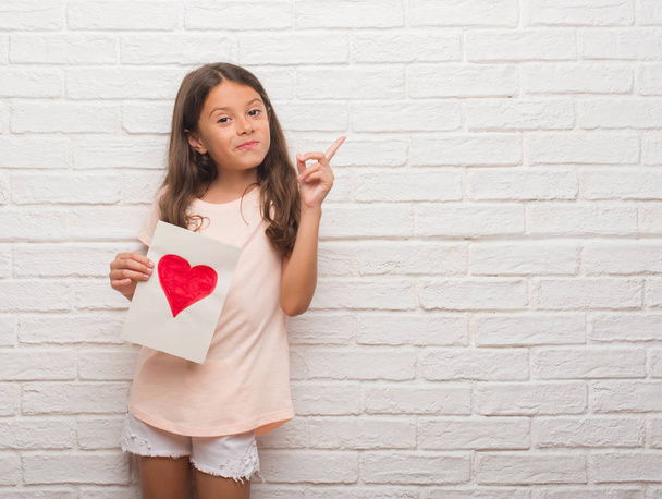 Jovem criança hispânica sobre parede de tijolo branco dando cartão mãe dia muito feliz apontando com a mão e o dedo para o lado
 - Foto, Imagem