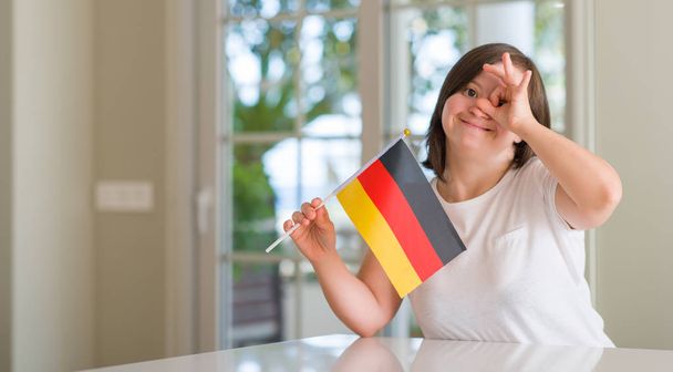 Σύνδρομο Down γυναίκα στο σπίτι κρατώντας την σημαία της Γερμανίας με χαρούμενο πρόσωπο χαμογελά κάνει εντάξει σημάδι με το χέρι στο μάτι ψάχνει μέσα από τα δάχτυλά - Φωτογραφία, εικόνα