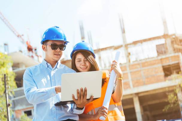 Asian man and woman engineer with the blue safety helmet meeting at the construction site with blueprints and laptop. - Фото, зображення