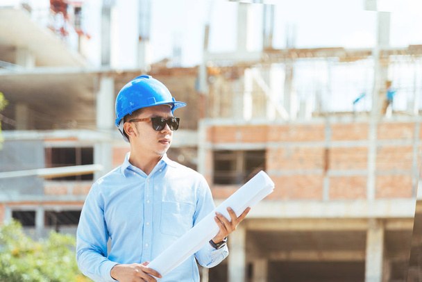 Civli engineer with blue safety helmet and sunglasses holding blueprint or drawing at construction site. - Фото, зображення