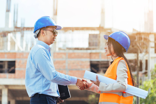 Close up Asian man civil engineer holding laptop and Asian woman architect holding drawing blueprint shaking hands at construction site. - 写真・画像