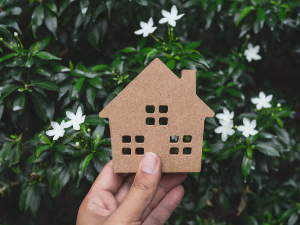 Mano sosteniendo casa de madera sobre hojas verdes y flores blancas de fondo. Concepto de ecología doméstica
. - Foto, Imagen