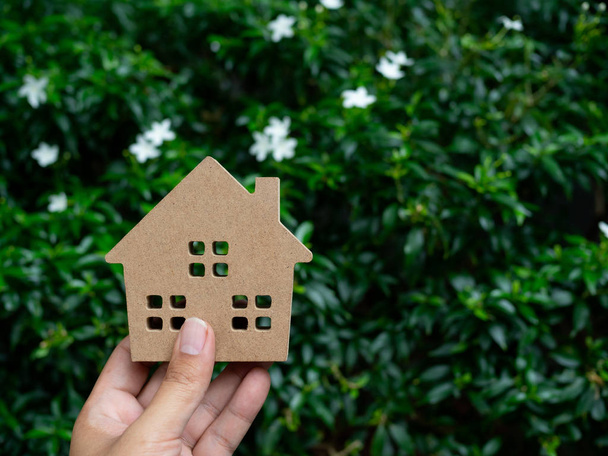 Mano sosteniendo casa de madera sobre hojas verdes y flores blancas de fondo con espacio de copia. Concepto de ecología doméstica
. - Foto, Imagen