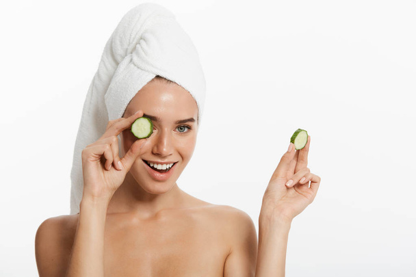 Close up beauty portrait of a smiling beautiful half naked woman with a towel wrapped around her hair holding cucumber slices and looking at camera isolated over white background - Photo, Image
