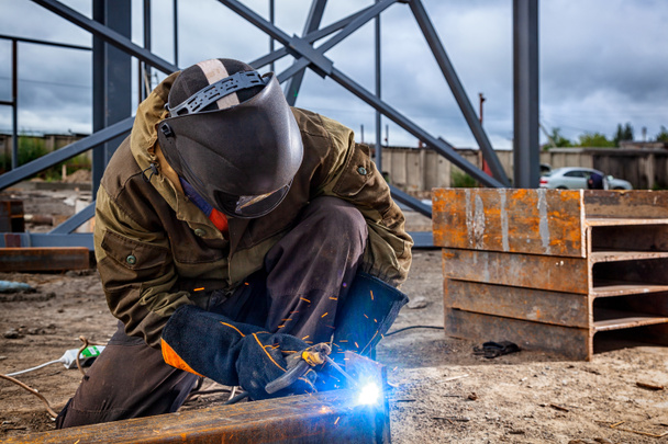 A young  man welder in brown uniform, welding mask and welders leathers, weld  metal  with a arc welding machine at the construction site, blue sparks fly to the sides - Photo, Image