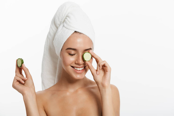 Close up beauty portrait of a smiling beautiful half naked woman with a towel wrapped around her hair holding cucumber slices and looking at camera isolated over white background - Φωτογραφία, εικόνα