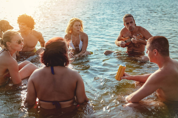 Groep gelukkige vrienden zitten in water en man ukulele spelen bij zonsondergang - Foto, afbeelding