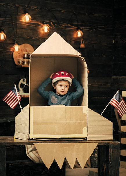 Boy play with rocket, cosmonaut sit in usa rocket made out of cardboard box. Kid in helmet sit in cardboard hand made rocket with usa flags. Child boy play american astronaut. Cosmonaut concept. - Zdjęcie, obraz
