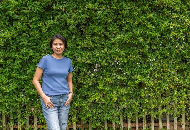 Vrouw poseren voor verlaat hek of hedge. Middelste leeftijd Aziatische vrouw permanent en glimlachen. - Foto, afbeelding