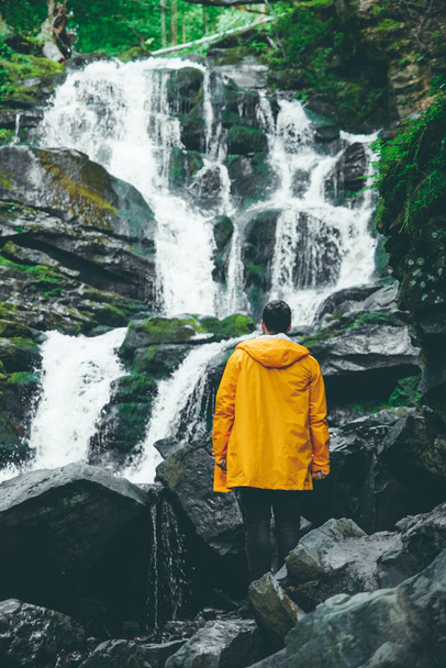 Mann steht in gelbem Regenmantel und blickt auf Wasserfall. Wanderkonzept - Foto, Bild