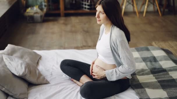Pregnant lady is caressing her tummy sitting on double bed at home and enjoying rest and leisure time. Modern interiors, expecting mothers and tenderness concept. - Filmati, video