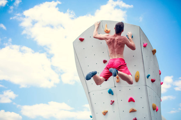 Photo from back of athlete guy in red shorts practicing on wall for climbing against blue sky with clouds - Photo, Image