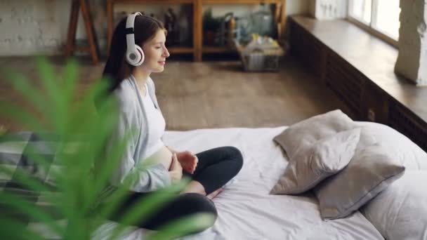 Cheerful young pregnant woman is stroking her belly and listening to music enjoying beautiful song wearing headphones and looking out of the window sitting on bed at home. - Filmagem, Vídeo