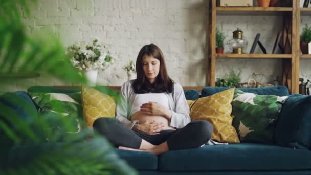Csinos barna terhes fiatal nő a Körvonalazás ő gyomor gondolkodás baba, ülő kanapé, modern apartman, sok növény és fából készült bútorok. - Felvétel, videó