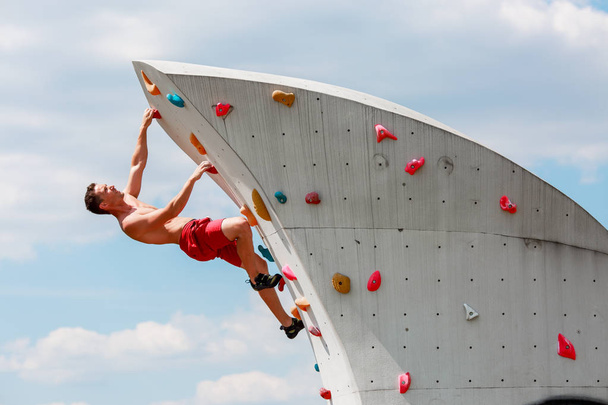 Photo of young sportsman in red shorts on wall for rock climbing against blue sky with clouds - Fotografie, Obrázek