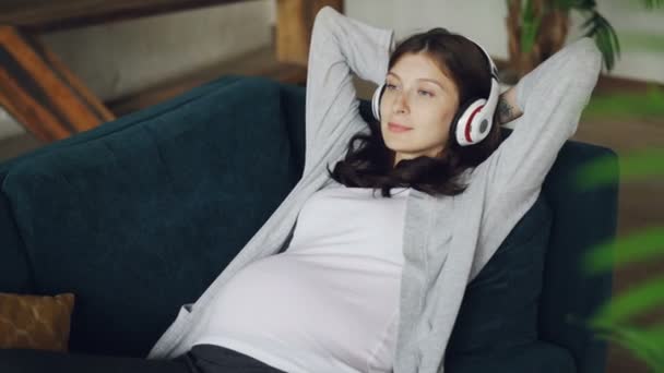 Beautiful girl expecting mother is listening to music wearing headphones and relaxing on sofa at home enjoying rest and songs. Family, relaxation and happy pregnancy concept. - Video, Çekim