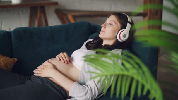 Pregnant young woman with dark hair is listening to music wearing modern wireless headphones and stroking her belly expressing love. Motherhood, technology and home concept. - Filmagem, Vídeo