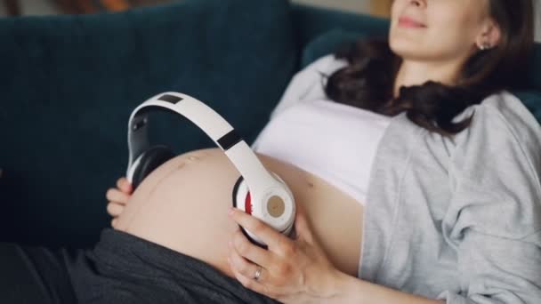 Smiling pregnant woman is lying on comfortable couch with headphones on her big tummy expressing love for her baby. Modern technology and motherhood concept. - Imágenes, Vídeo