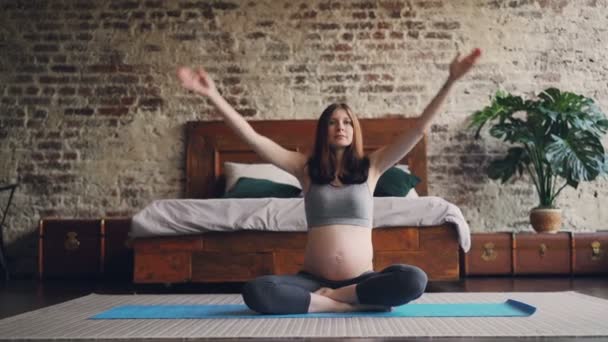 Sporty pregnant woman is practising yoga at home sitting on mat on bedroom floor raising hands, putting palms together then relaxing and breating in lotus position. - Video
