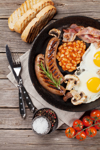 Anglická snídaně. Smažená vejce, párky, slanina, fazole, toasty, rajčata, pomerančová šťáva a šálek kávy na dřevěný stůl. Pohled shora - Fotografie, Obrázek