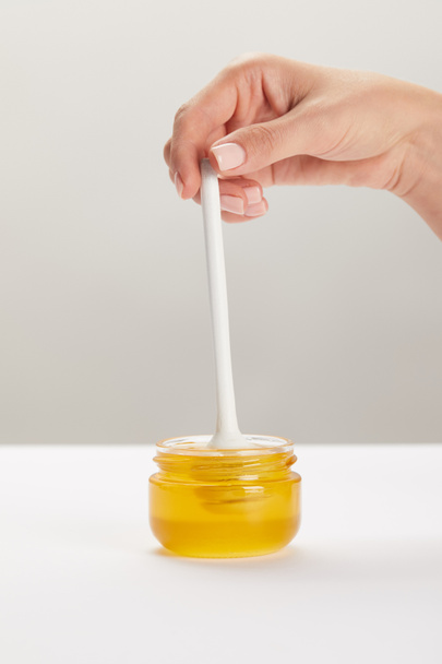 Μερική άποψη της γυναίκας κρατώντας ξύλινα βαθύτερα με γυάλινο βάζο γεμάτο μέλι μέλι σε λευκή επιφάνεια - Φωτογραφία, εικόνα