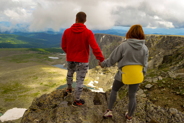 joven pareja amorosa de turistas tomados de la mano de pie en la cima de un acantilado y mirando un pintoresco valle de montañas, rocas, lagos y cielo azul con nubes blancas bajas. Sígueme por detrás.
 - Foto, imagen