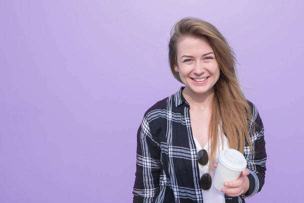 Šťastná dívka stojící na fialovém pozadí s šálkem kávy v její ruce úsměvy a dívá na kameru. Dívka s drinkem na pozadí fialové zdi - Fotografie, Obrázek